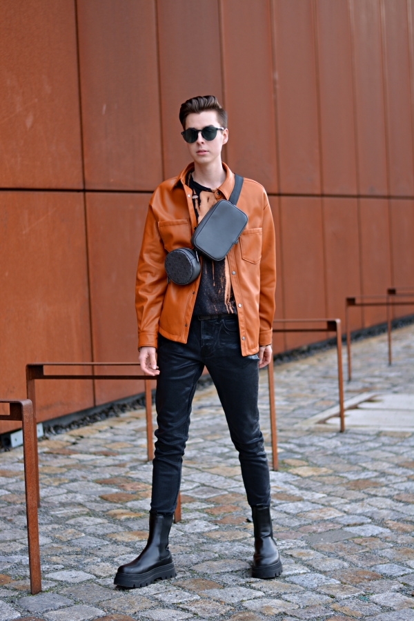 Modeblogger vor rostiger Wand in Overshirt in orange, mit grauer Bauchtasche und schwarzen Boots