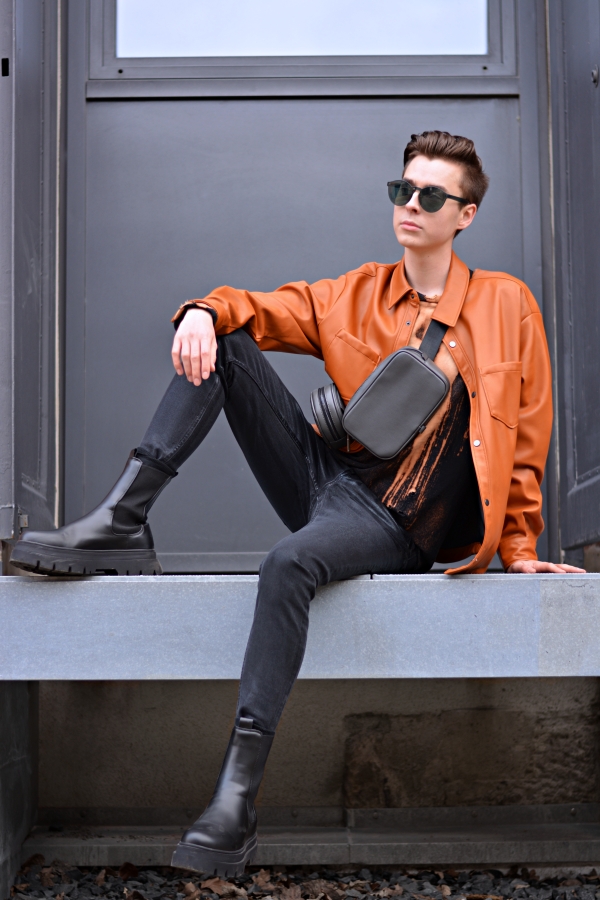 Junger Modeblogger sitzt auf Metallplattform. Er hat ein Overshirt in orange an und Boots