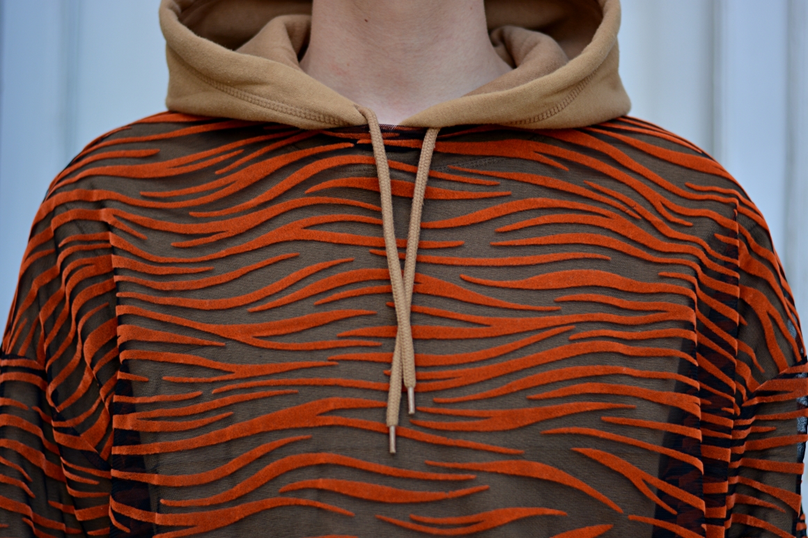 Detailbild von hellbraunem Hoodie darauf T-Shirt aus Mesh mit Tigerprint. 
